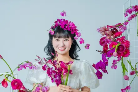 白いドレスと花の冠をつけた人物が鮮やかなピンク・紫の生花に囲まれ、花を手にして笑顔を向ける