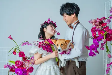 ピンクと紫の生花に囲まれ、花の冠をつけた白いドレスの人物と犬を抱いた人物が向き合う