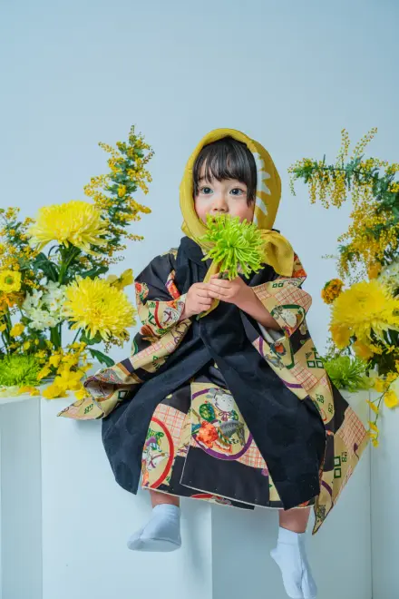 黄色いずきんと黒い絵柄着物を着て緑の菊を口に当て、黄色い花に囲まれた台に腰掛ける七五三3歳の子ども