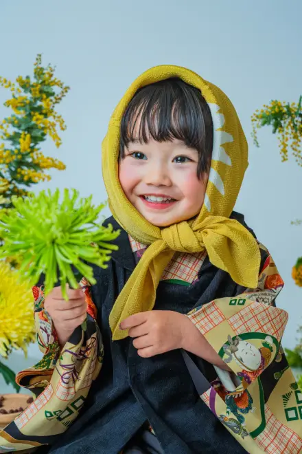 黄色いずきんと黒い絵柄着物で緑の菊を手に持ち、黄色い花々の前で満面の笑顔を向ける七五三3歳の子ども