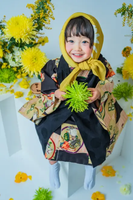 からし色のずきんに黒い絵柄着物を着て緑の菊を手に持ち、黄色の花びらが散る中で笑顔を見せる七五三3歳の子ども