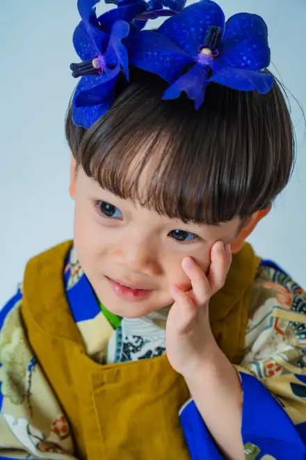 大きな青い胡蝶蘭の髪飾りをつけ、からし色と青の鮮やかな着物で頬に手を当ててこちらを見つめる七五三の子ども