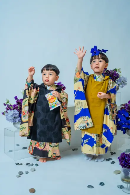 黒い着物とからし色・青の着物をそれぞれ着た2人の子どもが手を上げて、紫の花に囲まれて並ぶ七五三