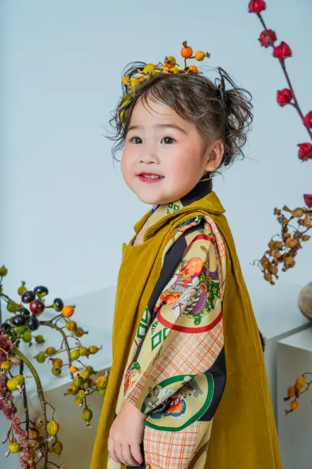 橙と黄の木の実の冠をつけ、からし色の着物でドライベリーの枝の前に横向きで立って微笑む七五三3歳の子ども