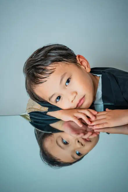 黒い羽織を着た子どもが反射するテーブルに頬と手を乗せ、鏡に映った自分と向き合うようにこちらを見つめる