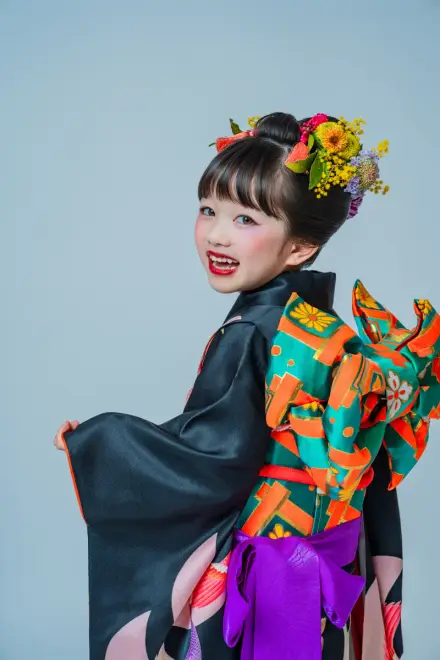 黒地の着物に鮮やかな緑とオレンジの帯を背に、花飾りの子どもが振り返って満面の笑顔を見せる
