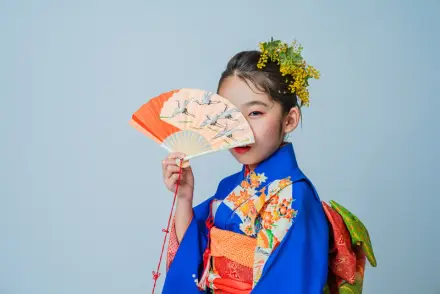 青い花柄着物を着てミモザの髪飾りをつけた七五三の女の子が、オレンジの扇子で顔を半分隠しながらこちらを見る