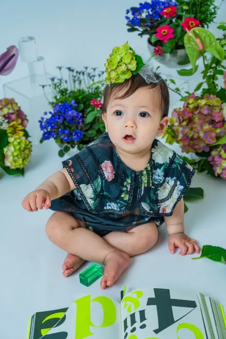 アジサイの髪飾りをつけた花柄ロンパースの赤ちゃんが、色とりどりの生花に囲まれてちょこんと座る