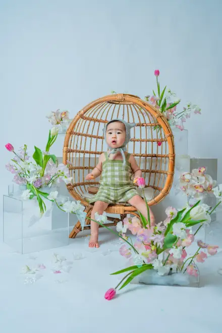 緑のチェック柄ロンパースを着た赤ちゃんが、丸いラタンチェアに腰かけ、チューリップや白い花々に囲まれる