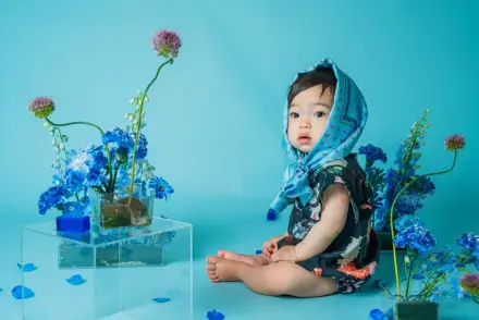 青いスカーフを頭に巻いた赤ちゃんが、水色の背景と青い生花に囲まれてこちらを振り返る
