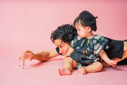 ピンク背景で大人が床に伏せてグラスに手を伸ばし、背中にベレー帽の赤ちゃんが乗って覗き込む