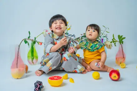 花柄の服を着た子どもとオレンジ色の服の赤ちゃんが、グレープフルーツやレモンなどの果物と花瓶に囲まれて並んで座る