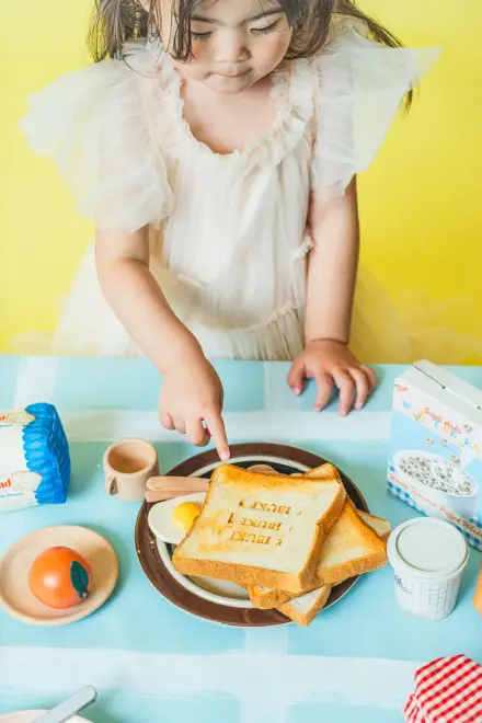 白いフリルドレスの子どもが青いテーブルのトーストを指差し、おもちゃ食材が並ぶ黄色い背景の前で見下ろす