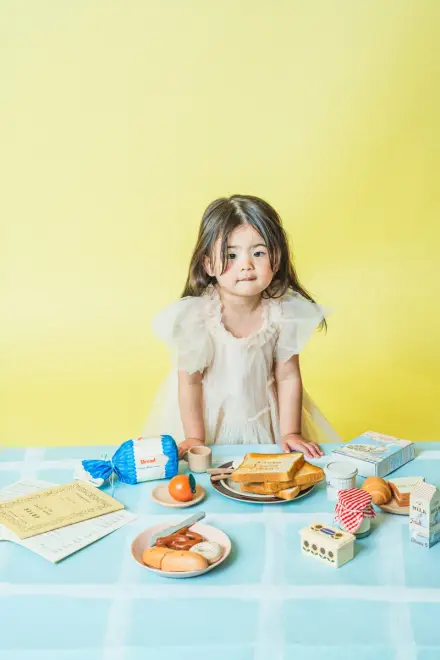 白いフリルドレスの子どもが青いチェックテーブルの前に立ち、パンや食品小物が広がる黄色い壁の前でこちらを見る