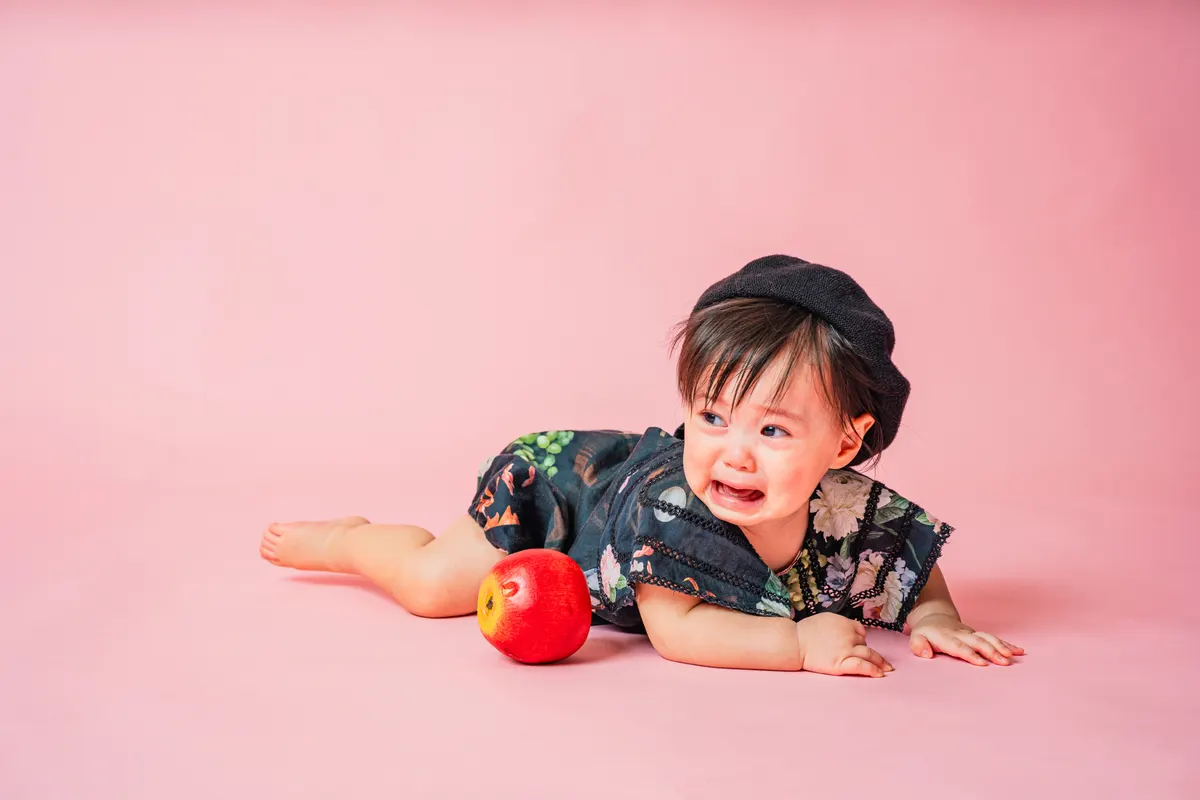 ピンク背景でベレー帽をかぶり林檎とうつ伏せになる赤ちゃん（Classic Portrait シーン）