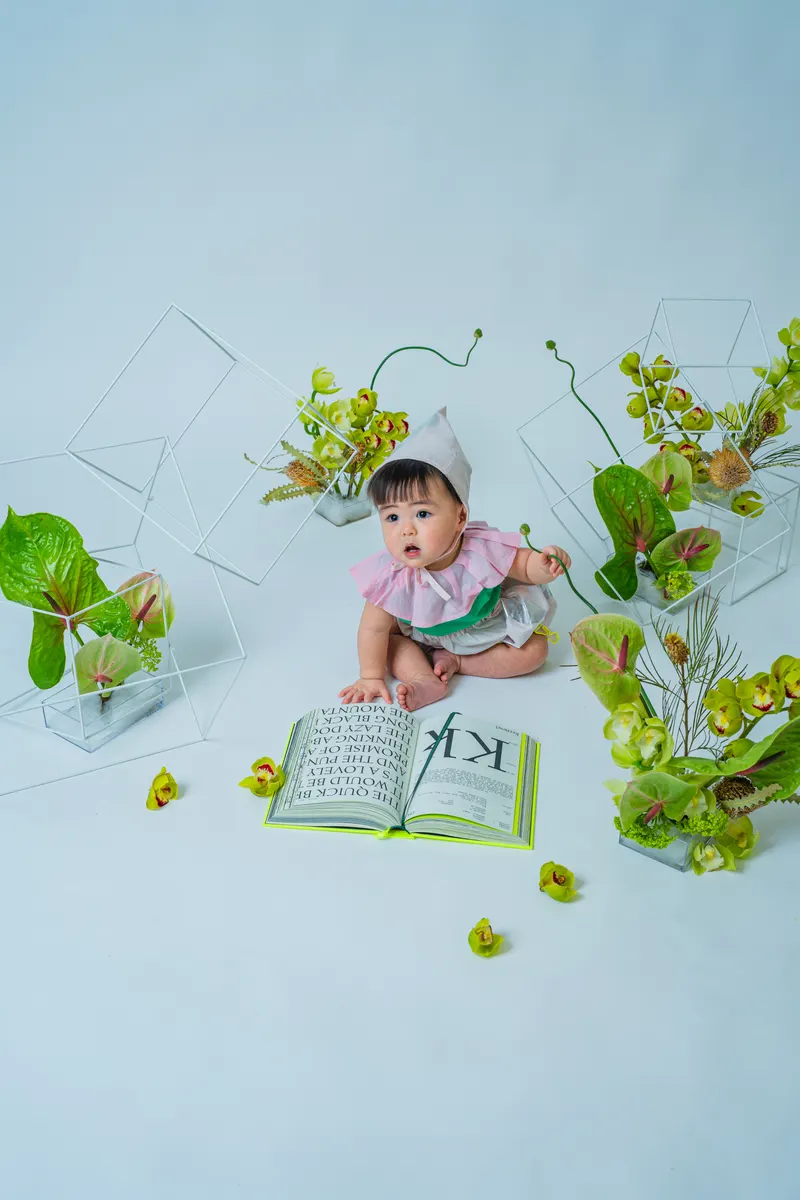 開いた本と緑の花に囲まれて座る赤ちゃん（Garden シーン）