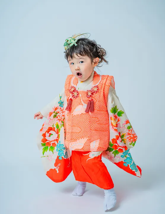 白地に牡丹や鶴の柄、朱色の被布を合わせた着物「芍薬」を着た女の子