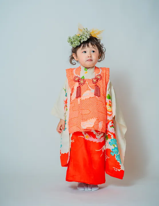 白地に牡丹や鶴の柄、朱色の被布を合わせた着物「芍薬」を着た女の子