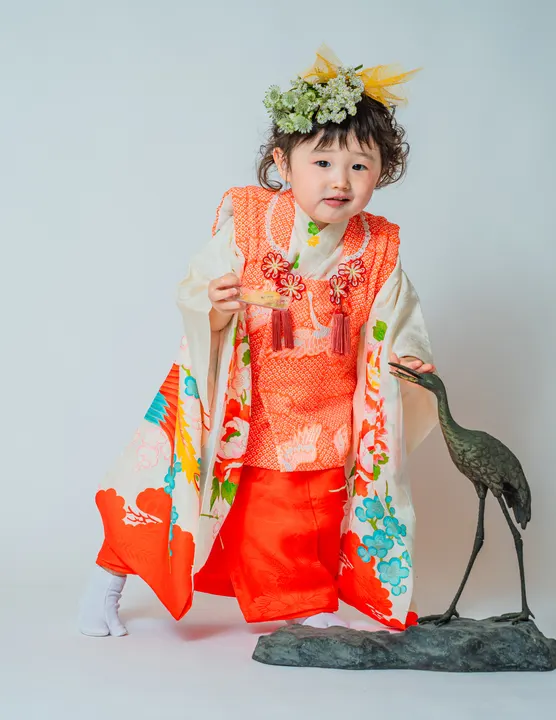 白地に牡丹や鶴の柄、朱色の被布を合わせた着物「芍薬」を着た女の子