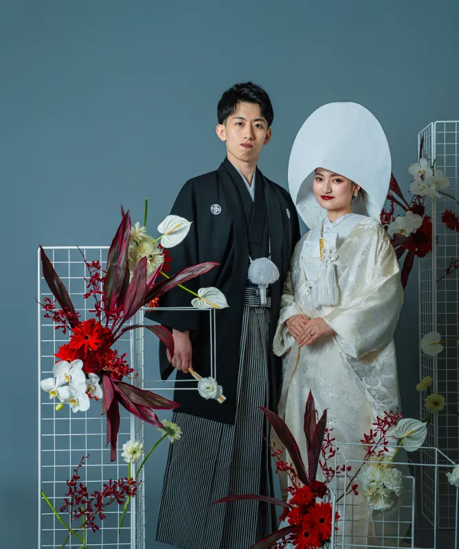 赤い装花を背に並ぶ黒紋付袴の新郎と綿帽子に白無垢の花嫁(和装シーン)
