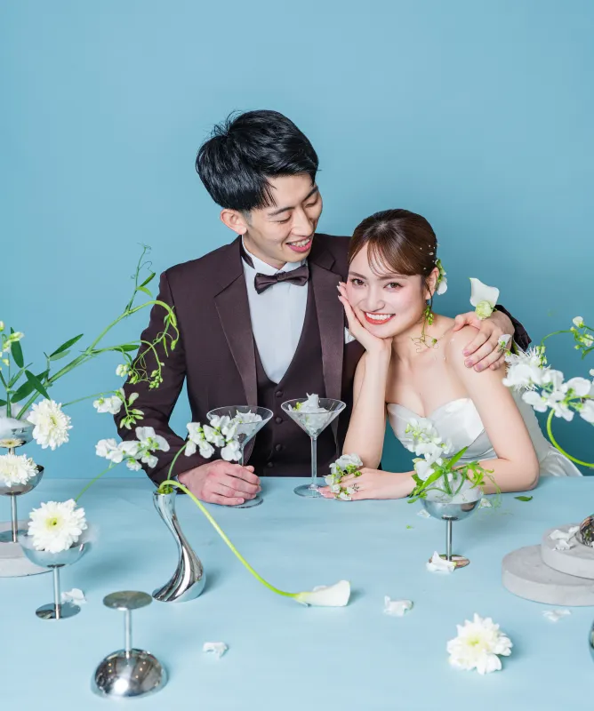 白い装花のテーブルで頬杖をつく花嫁に寄り添う新郎(Table シーン)